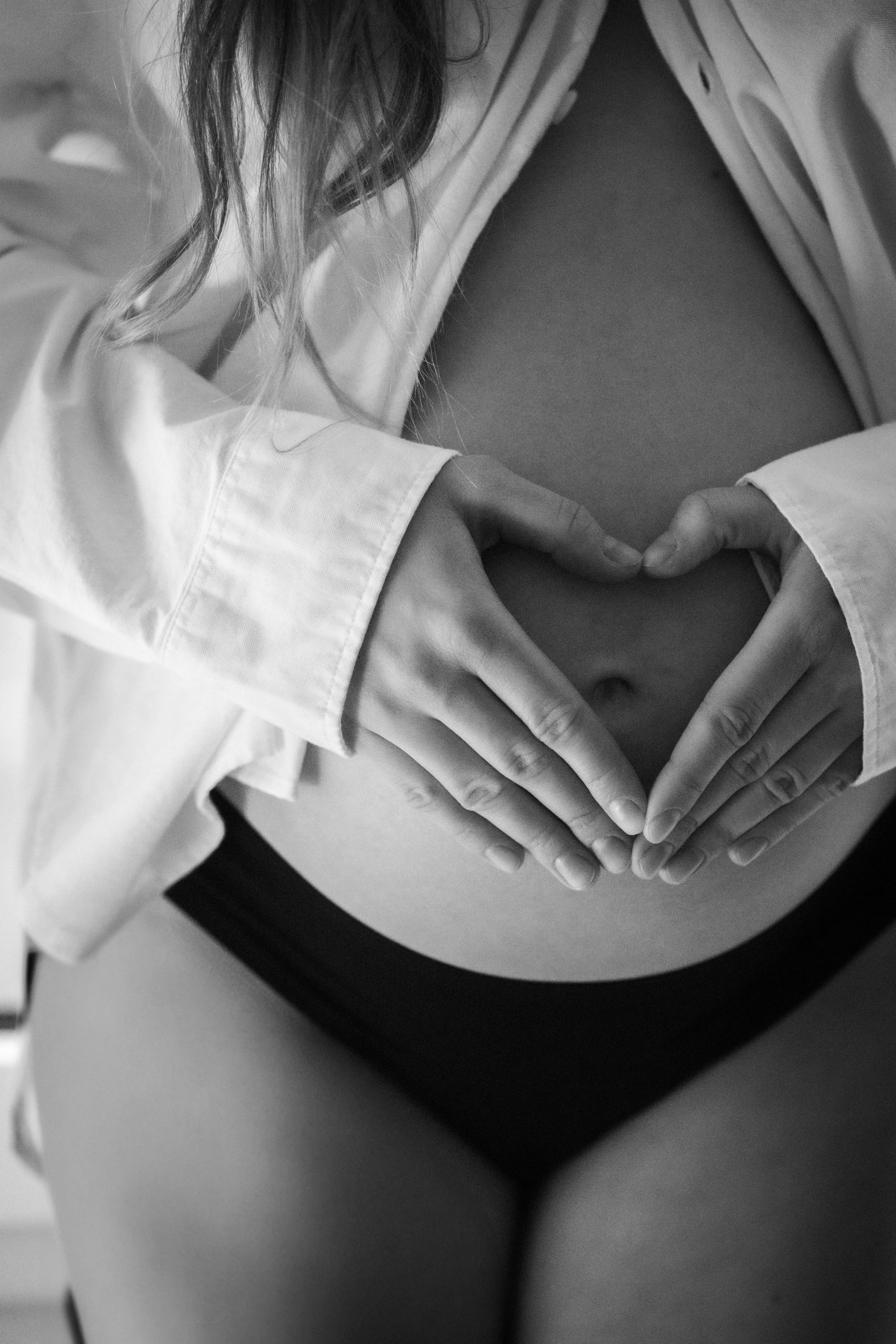 Black and white image of a pregnant woman forming a heart shape on her belly with her hands.
